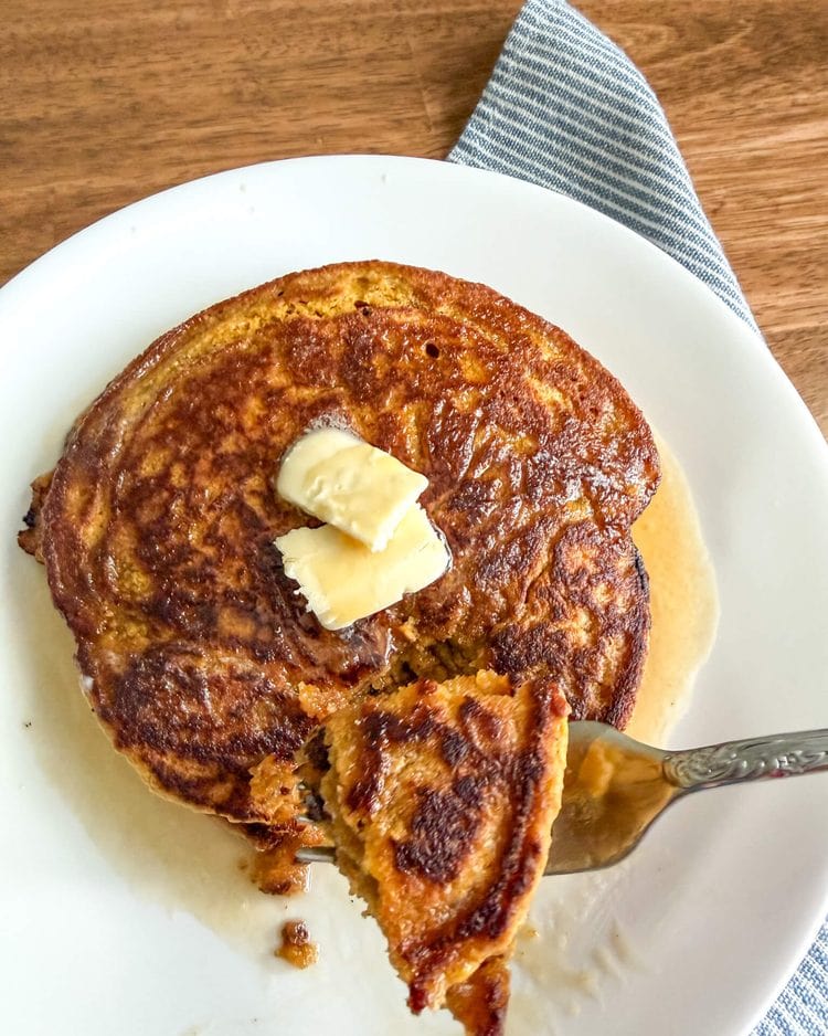 Pumpkin pancakes with fresh milled flour with a bite taken out on a fork. Pancakes are on a white plate on a wooden table with a blue and white striped cloth napkin next to them.