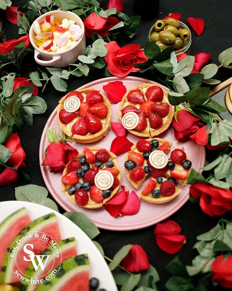 Beauty and the Beast Party table ideas. A dark tablecloth covered in Paul bakery strawberry tarts studded with berries. The plate wrapped in a rose garland and covered in rose petals. 