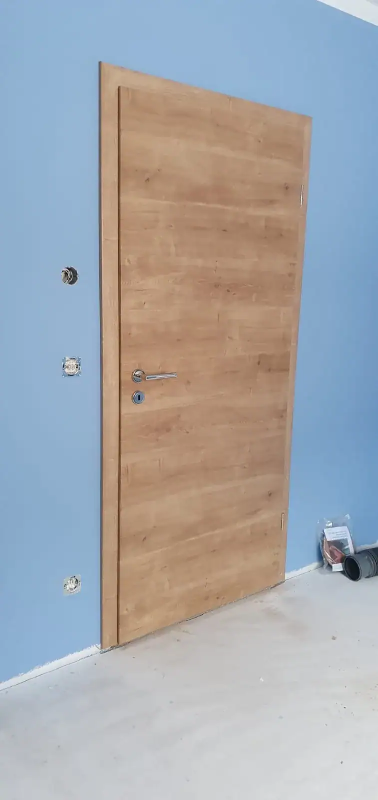 Zimmertür von Fütterer Fenster & Türen GmbH mit hellem Holzdekor und modernem silbernem Türgriff sowie Schloss. Die Tür ist in eine hellblaue Wand eingebaut und zeigt eine schlichte, elegante Optik in einem Raum, der sich im Ausbau befindet.