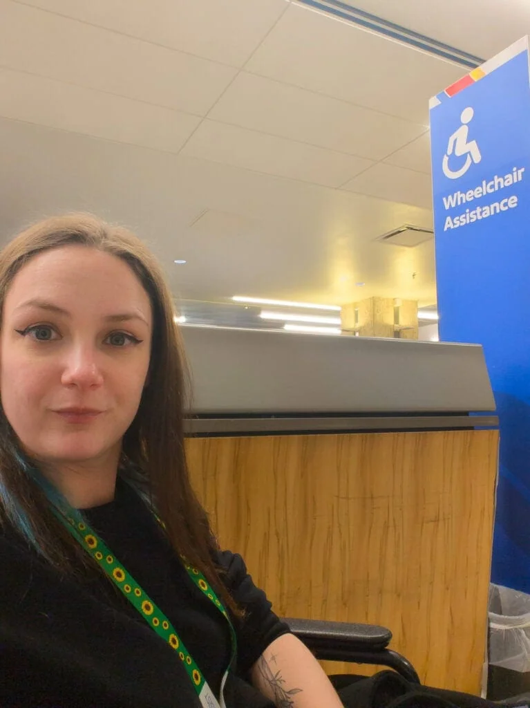Woman sitting in an airport wheelchair near a counter with a blue sign that reads “Wheelchair Assistance” while waiting for Southwest airport assistance.