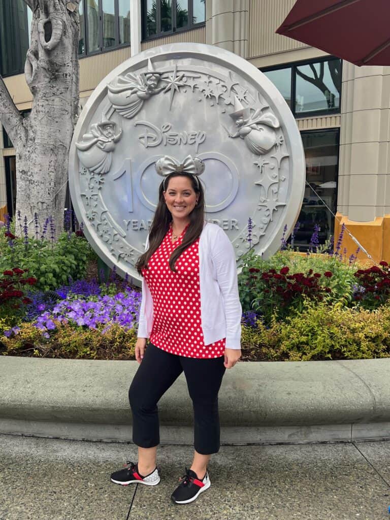 Disney 100th anniversary celebration outside Disney park, woman smiling in red polka dot top, Minnie Mouse ears, surrounded by flowers.
