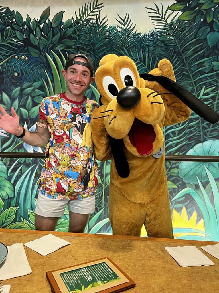Smiling man with Disney character Pluto in jungle-themed background at 407 and Beyond Vacation Co.