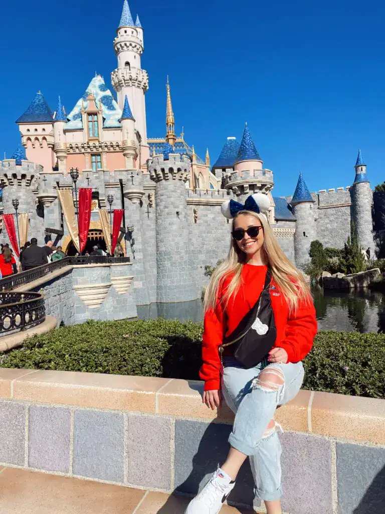 Colorful Disney castle skyline at Disneyland with a smiling woman wearing Minnie Mouse ears and sunglasses in front.