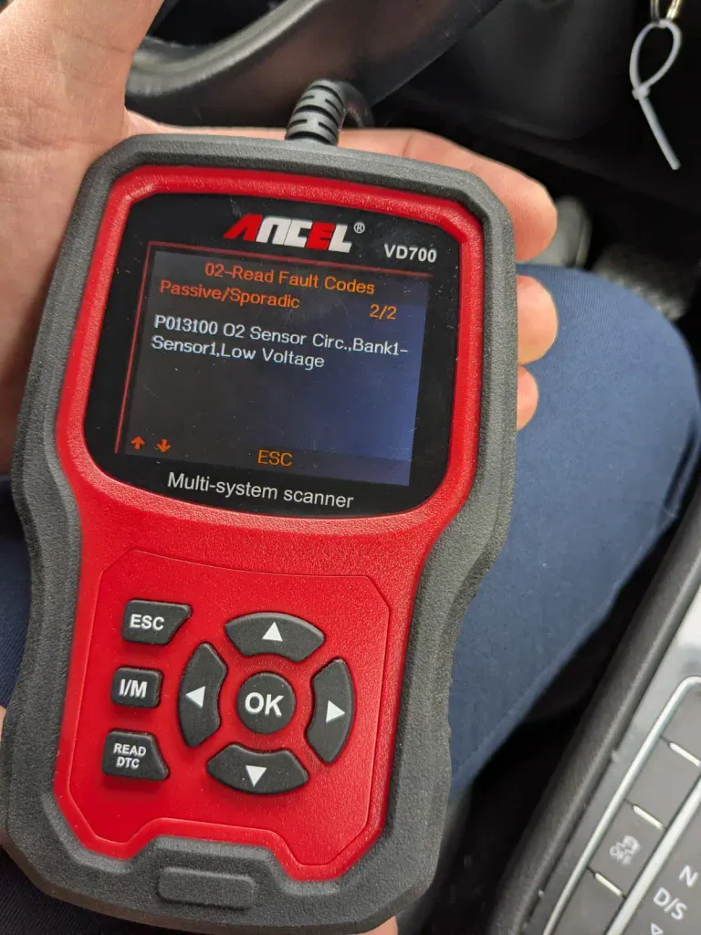 Close-up of a red and black ANCEL VD700 multi-system OBD scanner held in a car, screen displaying fault code P013100 O2 Sensor Circ. Bank1-Sensor1 Low Voltage, 2 of 2 codes.