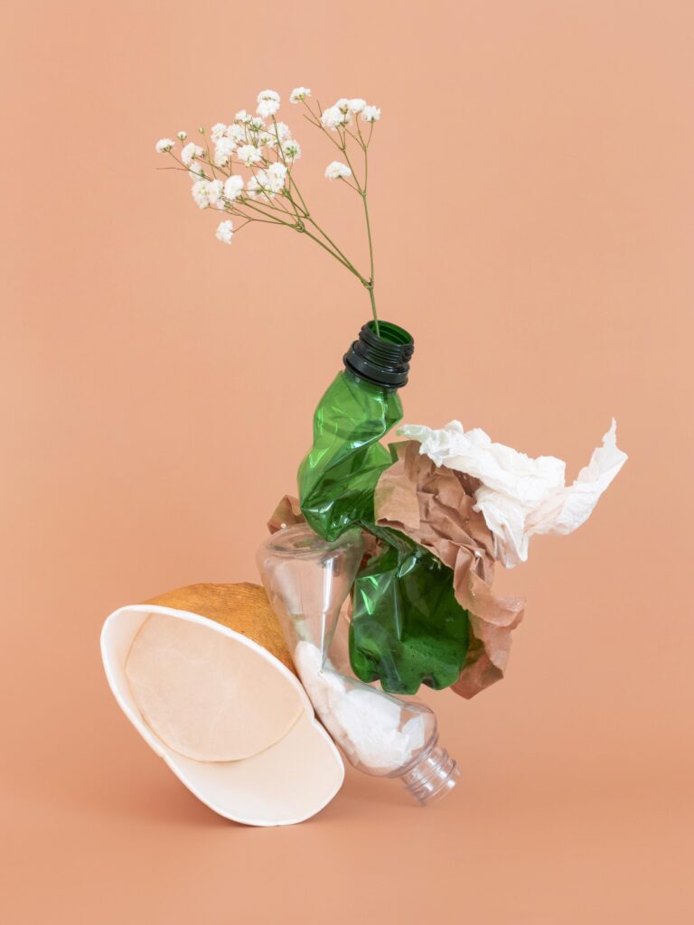 Composition artistique de déchets non écologiques : une bouteille en plastique verte froissée servant de vase pour une branche de gypsophile blanche, un gobelet en carton, une bouteille de lessive vide, des papiers froissés et un tissu blanc, sur un fond uni pêche.