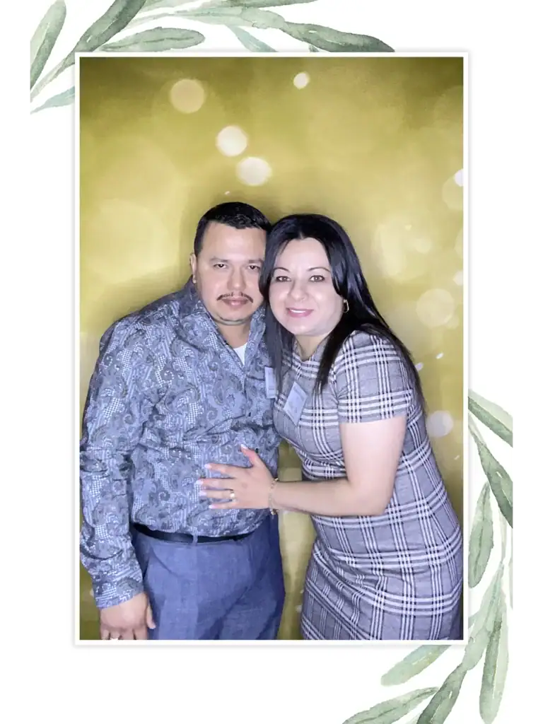 A couple poses in front of a golden bokeh-style backdrop, smiling together. The woman wears a plaid dress, and the man wears a paisley shirt.