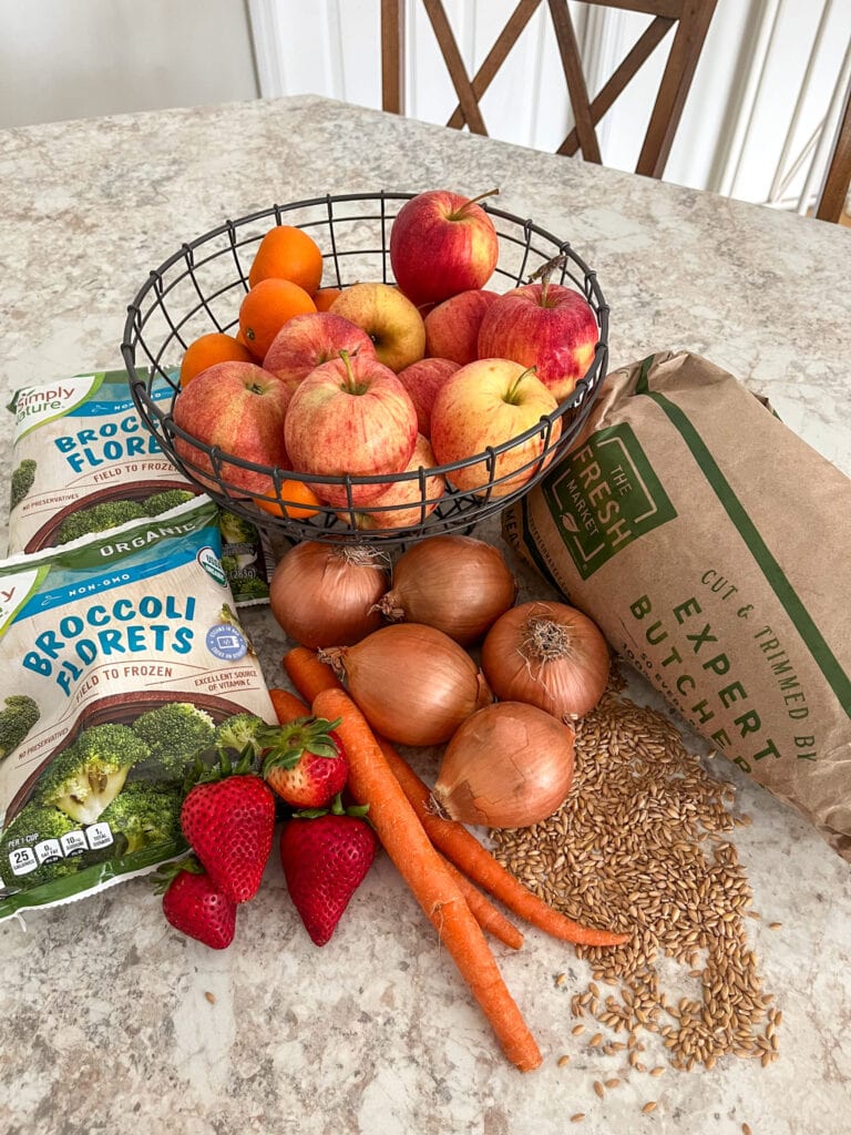Healthy food laying on a neutral colored kitchen countertop: frozen brocolli in bags, strawberries, carrots, onions, wheat berries, a roast in butcher paper, and oranges and apples in a fruit basket. 