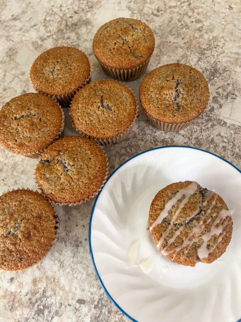 Blueberry muffin drizzles with powdered sugar icing on a white plate surrounded by other blueberry muffins on top of a kitchen countertop.