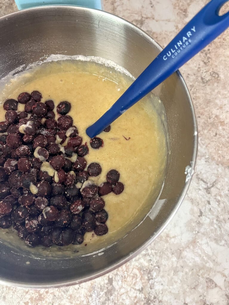 Folding blueberries in muffin batter om a stainless steel mixing bowl with a blue spatula.
