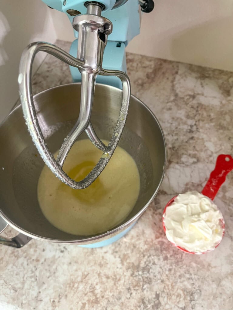 Wet ingredients for blueberry muffins inside a kitchen aid mixer with sour cream in a red measuring cup next to it.