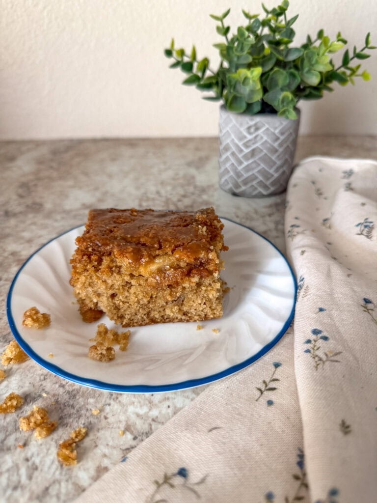 apple cake on plate on top of a neutral colored kitchen counter with a floral kitchen towel next to it