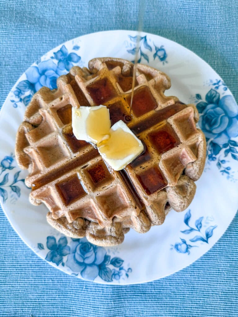 Einkorn waffles with two pats of butter and a stream of maple syrup being poured. Waffles are on a white plate with blue florals on a blue placemat.