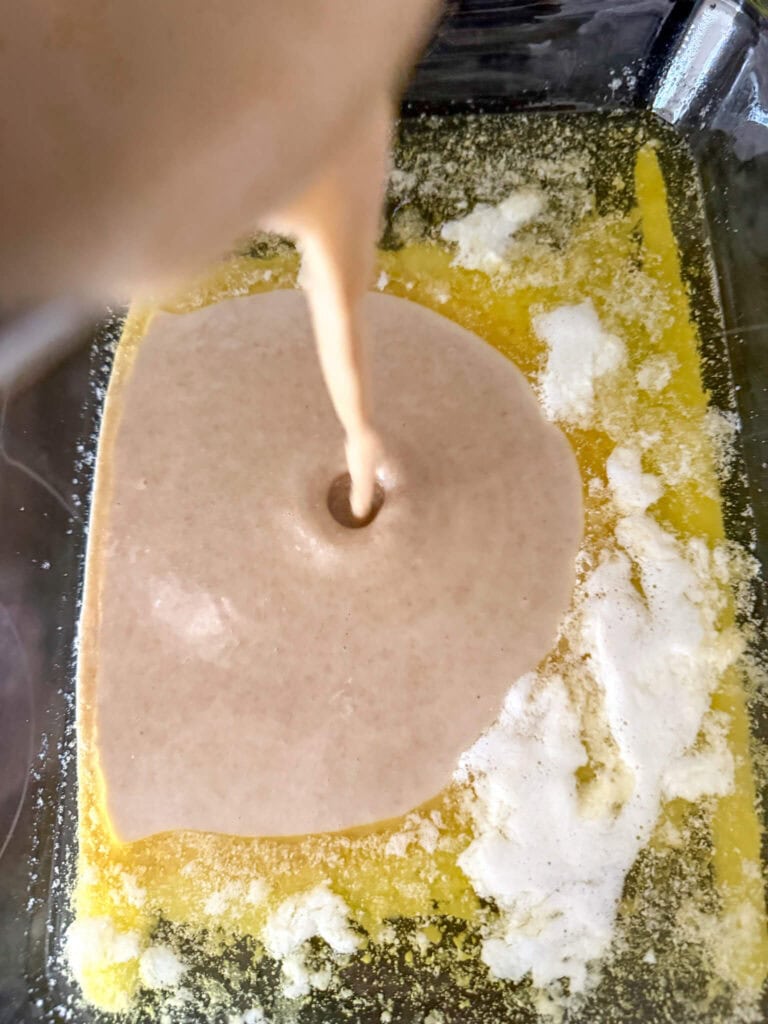 peach cobbler with fresh milled flour batter being poured into a glass baking dish containing melted butter