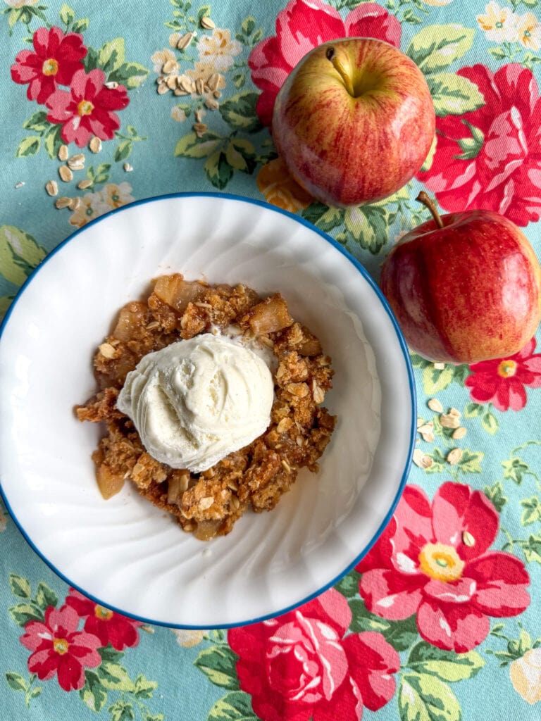 Bowl of fresh milled flour apple crisp on a bright colored kitchen placemat with two apples on the side.
