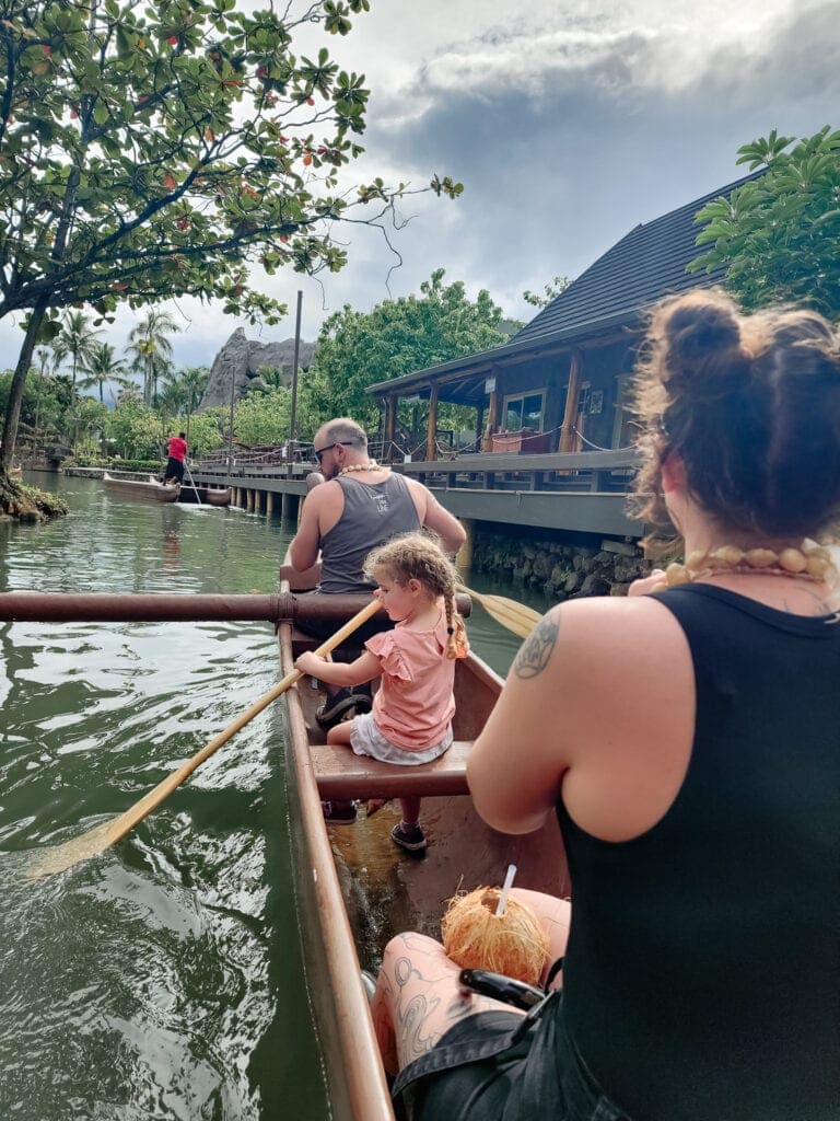 A family paddles a wooden outrigger canoe through a tranquil lagoon, passing Polynesian-style buildings and lush tropical trees, with the little girl helping row with her own paddle.