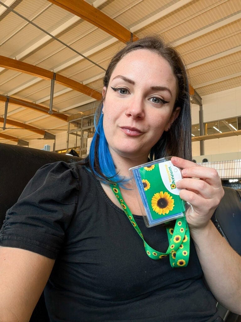 Phoenyx sits at an airport gate wearing a sunflower lanyard and holding up a matching Sunflower ID card, representing invisible disabilities.