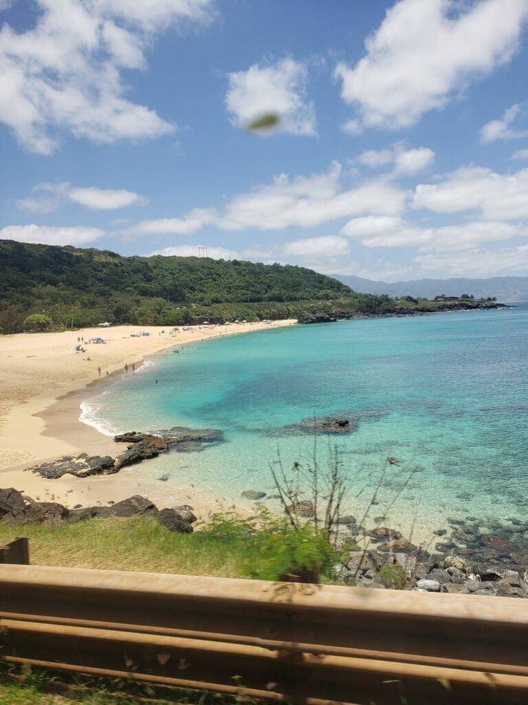 Scenic view of Kailua Beach on Oʻahu, Hawaiʻi, showing soft golden sand, clear turquoise water, rocky shoreline, and lush green hills under a blue sky with clouds.