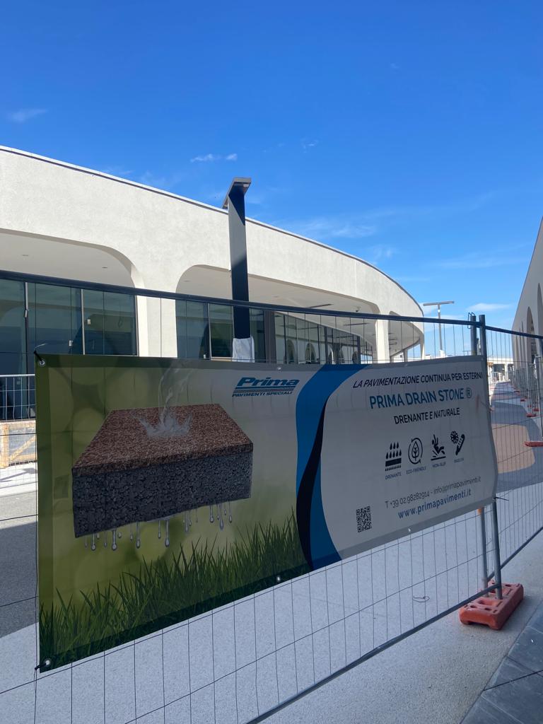 Area di collegamento tra padiglioni del centro commerciale TO.DREAM con pavimentazione drenante Prima Drain Stone®: il nostro cartello cantiere che illustra la soluzione drenante