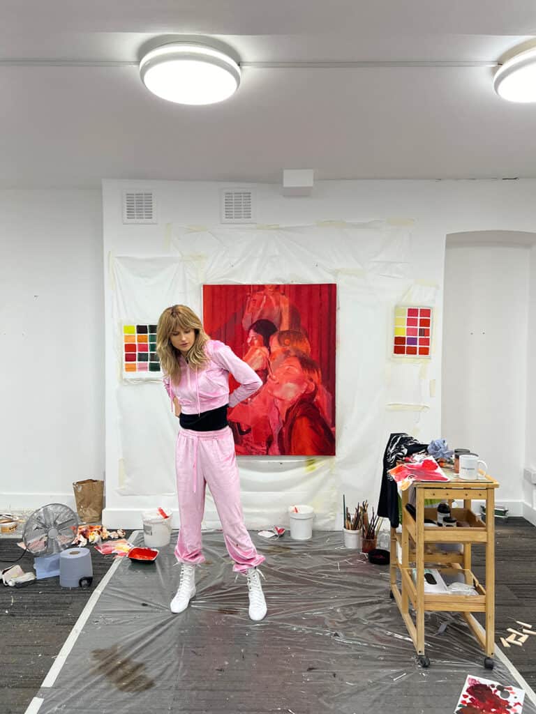 Artist studio scene from Nana Wolke’s “Nothing Left to Want” project, showing Nana in front of a large red painting surrounded by paints and materials.
