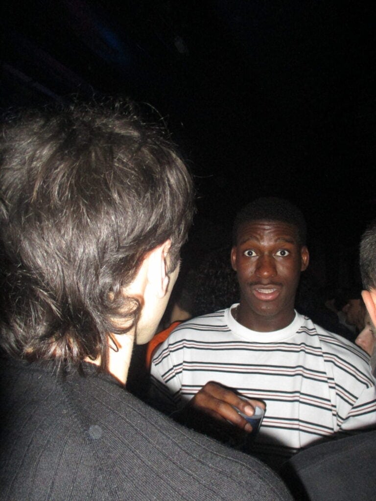 Two people are talking in a dimly lit nightclub at the 2much Festival. One person, facing the camera and holding a drink, looks surprised, while the other is seen from behind with only the back of their head visible.
