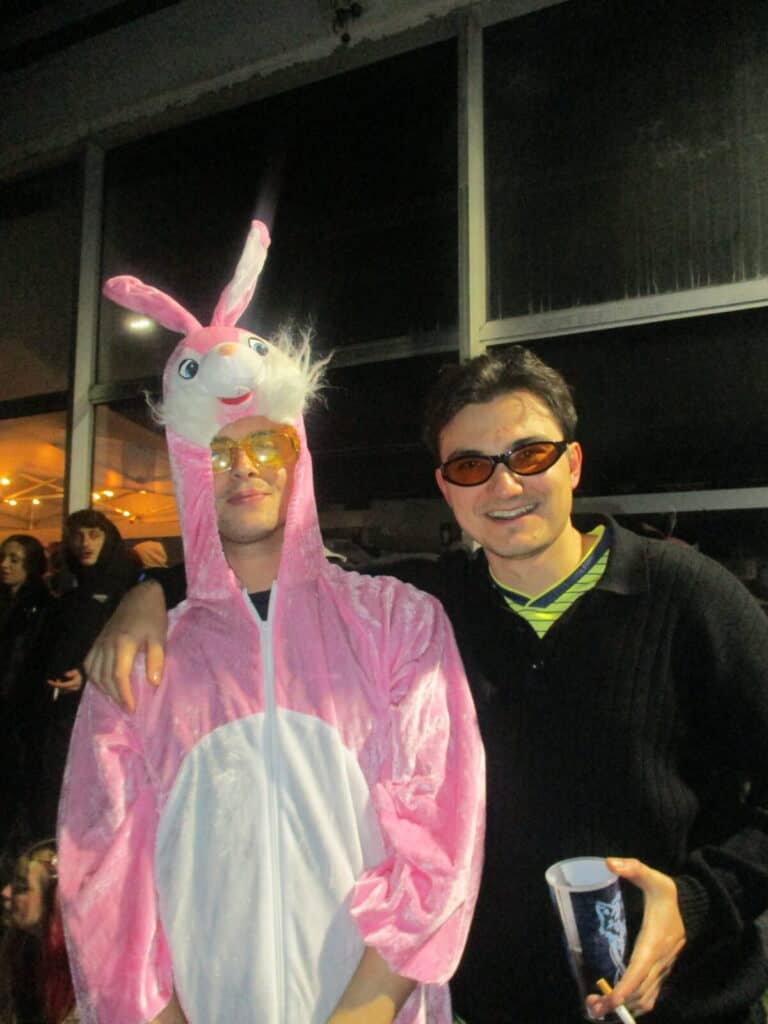 Two people posing together at 2much Festival—one in a pink bunny costume with a rabbit head hood and sunglasses, the other in dark clothing with orange sunglasses, smiling and holding a cup. People and lights fill the background.