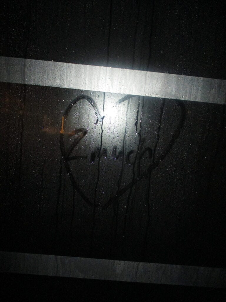A foggy window with a heart shape and the word Amor drawn on it captures the spirit of 2much Festival. Two horizontal white stripes cross the glass, while a bright light from behind highlights the heartfelt drawing.
