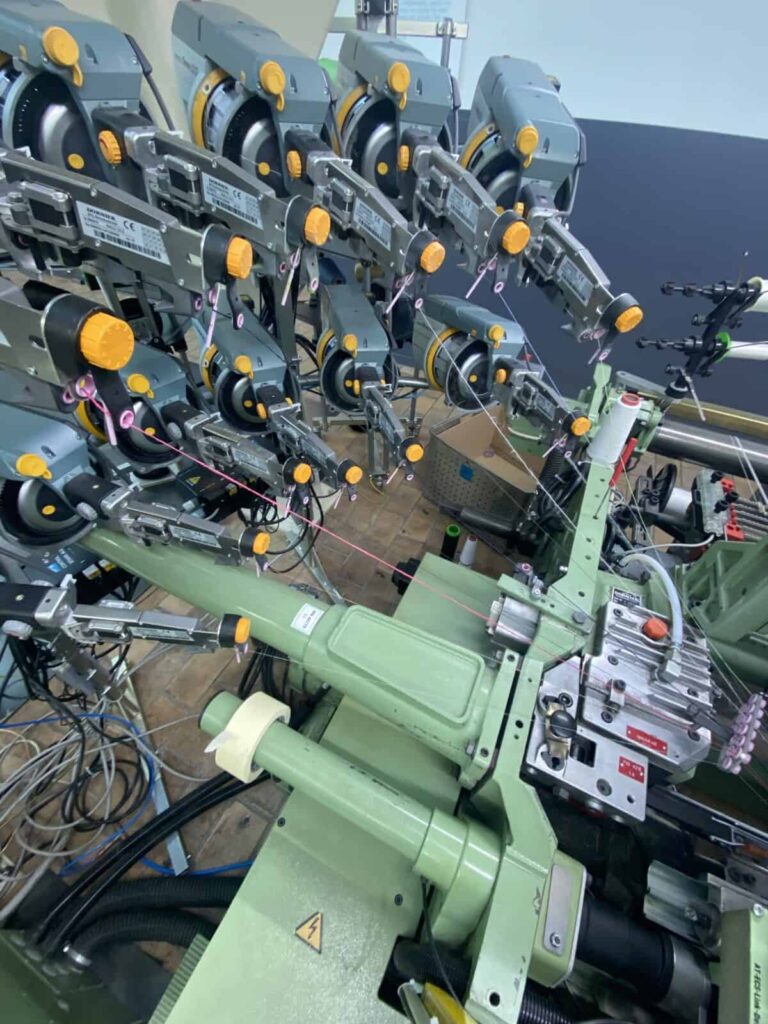 Close-up of a complex industrial weaving or textile machine with multiple spools, threads, gears, and metal components, showing intricate threading and machinery parts in operation.