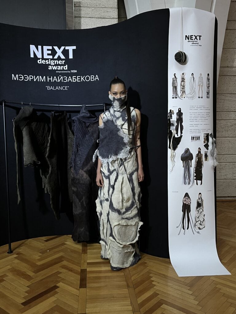 Meerim Naizabekova’s exhibition booth at Visa Fashion Week Almaty, displaying a white and grey felted marble-effect gown next to a vertical NEXT Designer Award moodboard.