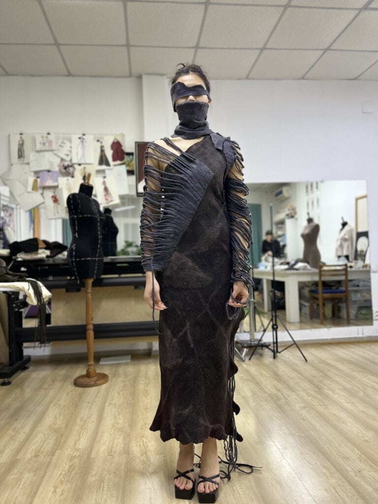 A full-length view of a model in Meerim Naizabekova’s "Balance" collection, showcasing a dark felted gown with horizontal fiber strands and a matching blindfold and mask.
