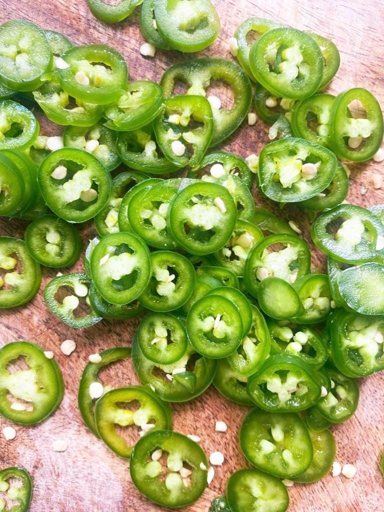 Top down image of a wooden board topped with sliced green Serrano peppers.
