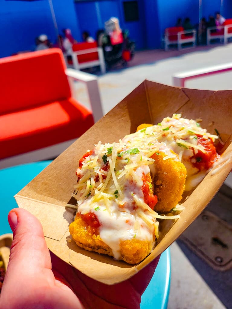 A handheld cardboard tray features golden-fried 'Impossible Chicken Parm Bites' topped with a rich tomato sauce and shredded cheese, all in sharp focus with a blurred outdoor seating area in the background.