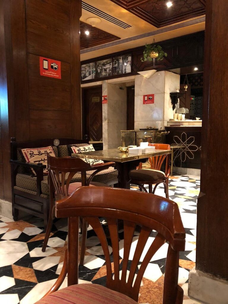 Interior shot of Khan El Khalili Café in Old Cairo, featuring traditional wood mashrabiya, brass lanterns, and guests enjoying shisha in a historic ambiance. Description: