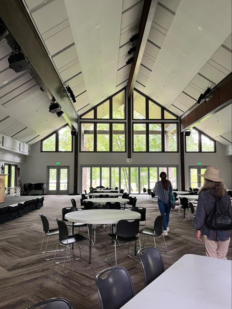 The main hall of the camp. Carpeted and filled with round tables. The back wall of the hall is entirely windows looking out onto natural grounds.