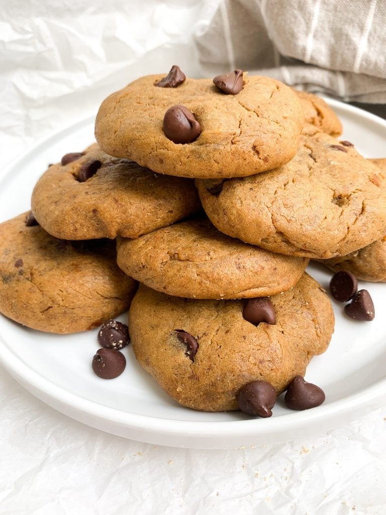 Chocolate chip protein cookies mounded on a white plate next to extra chocolate chips