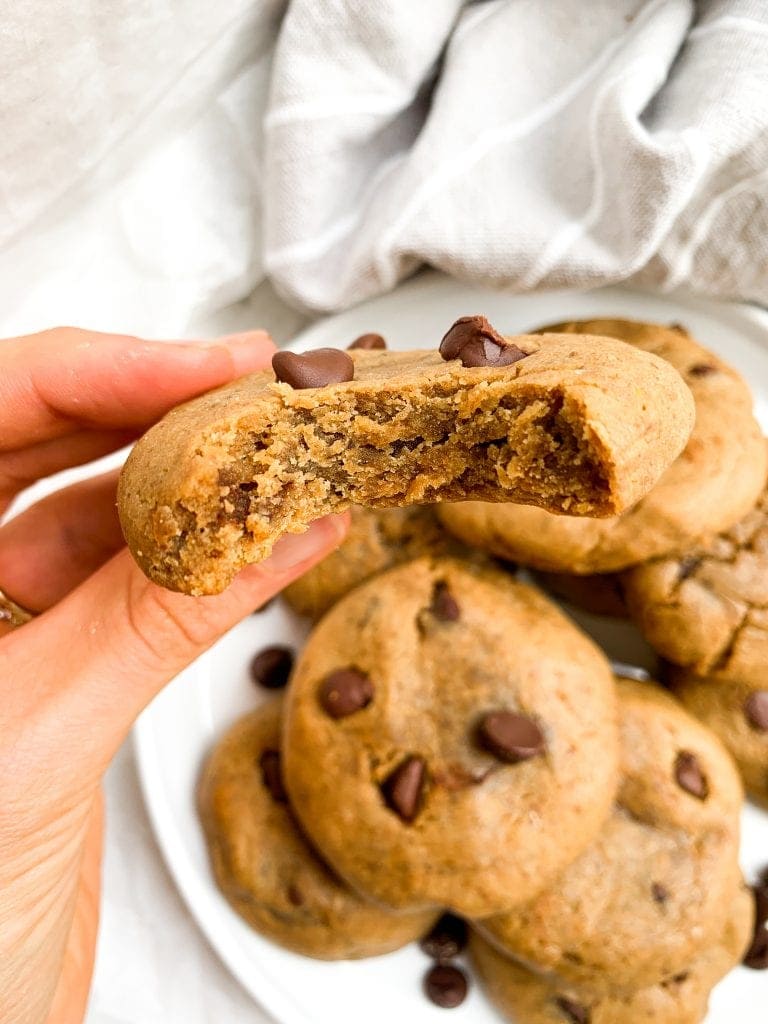 a plate of gluten free chocolate chip protein cookies in the background with a person holding one after taking a bite