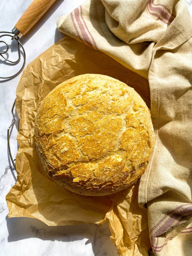 no knead bread, under parchment paper, wire rack, towel and on the side a bread a dough whisk, on marble background