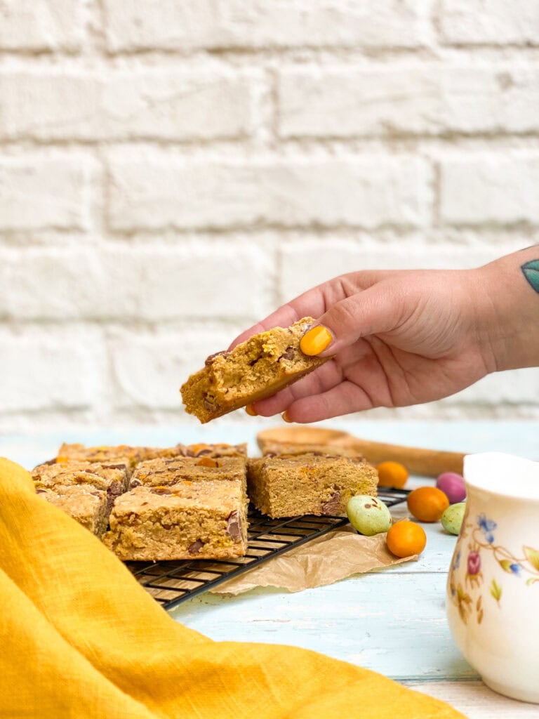 Super Easy Mini Eggs Blondies - A 30-minute Easter Traybake 2 mini eggs blondies on a cookie rack, mini eggs on the side, a hand reaching for one
