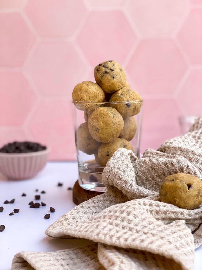 cookie dough bits in a clear glass, towel and choc chip in a bowl in the back, pink background