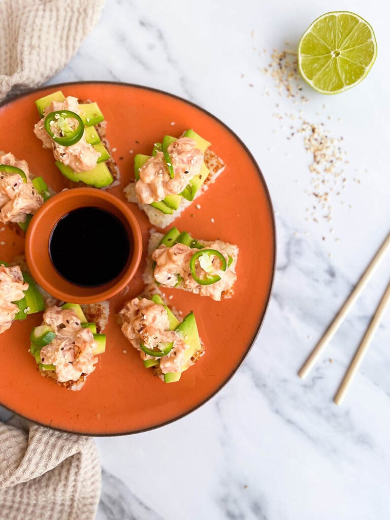orange dish with a bowl of soy sauce and an arrangement of crispy rice with spicy salmon, jalapeno and sesame seeds surrounded by wooden chopsticks and a lime