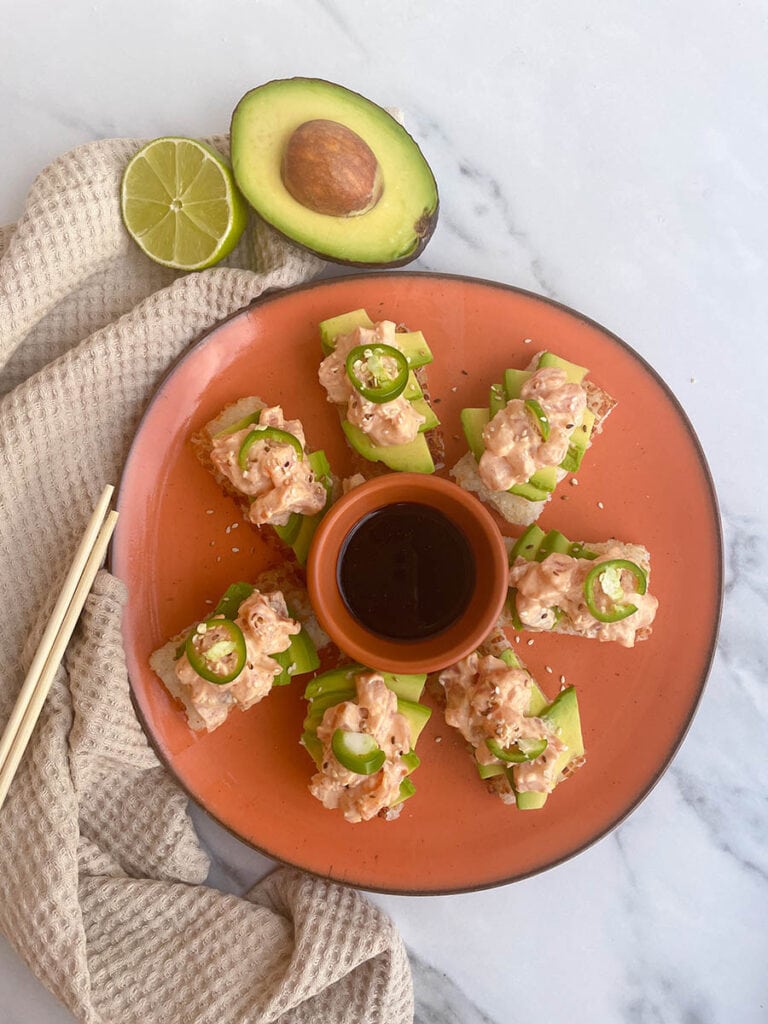 orange dish with a bowl of soy sauce and an arrangement of crispy rice with spicy salmon, jalapeno and sesame seeds surrounded by wooden chopsticks and a lime