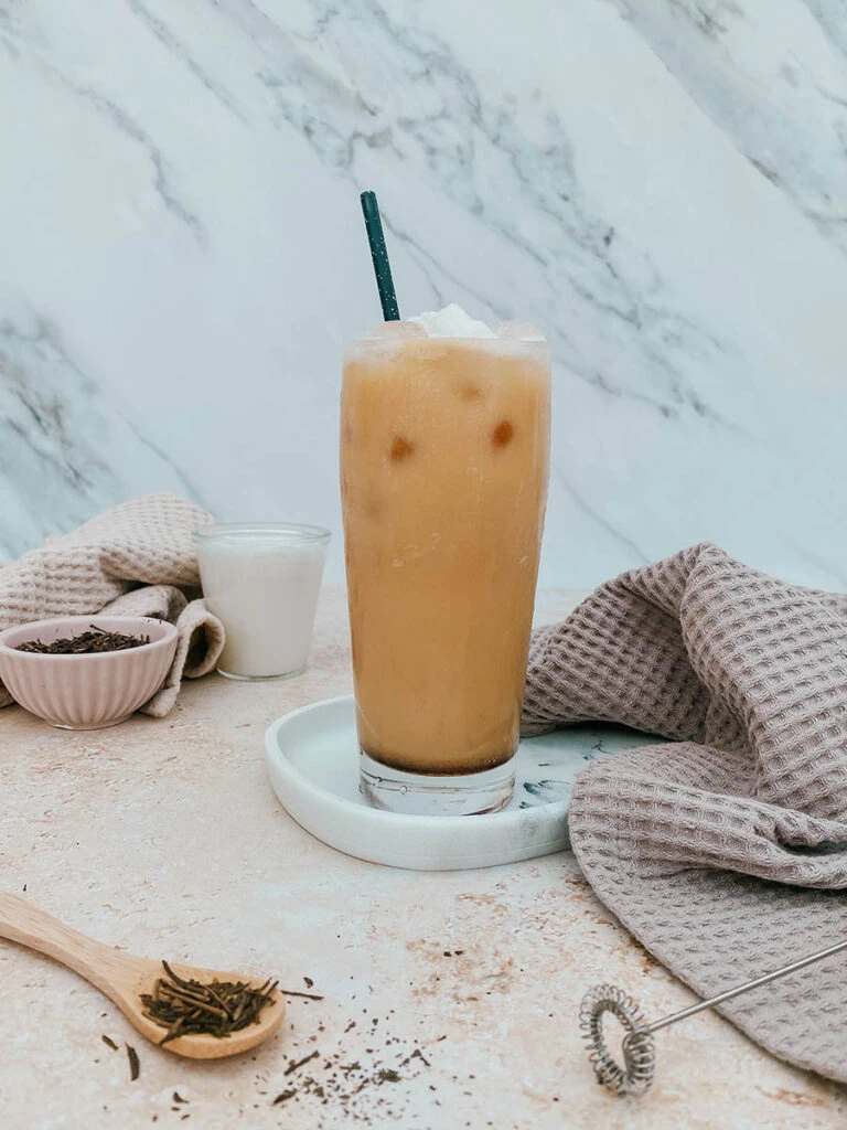 glass of iced hojicha latte, plus a glass of milk and a small pink bowl with tea leaves on the background