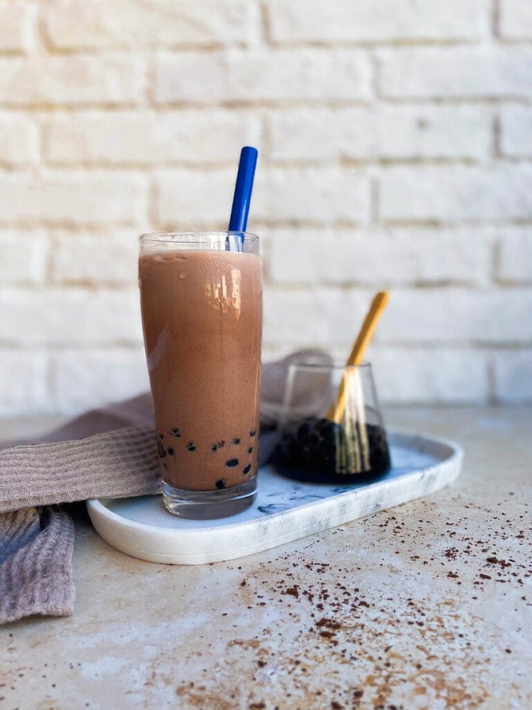 glass of chocolate milk tea, boba pearls in a glass container behind, towel and chocolate powder