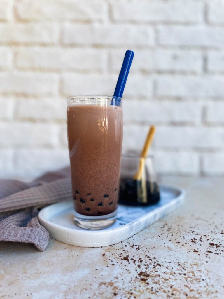 glass of chocolate milk tea, boba pearls in a glass container behind, towel and marble tray under