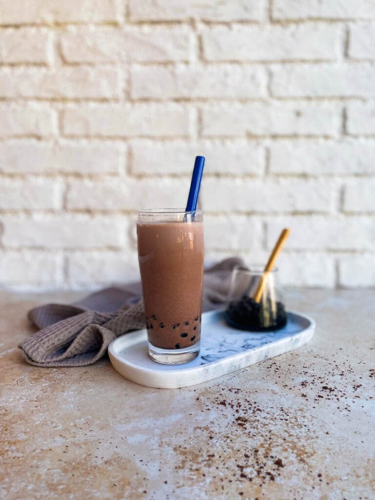chocolate milk tea, boba pearls in a glass container behind, towel, cocoa powder on the side