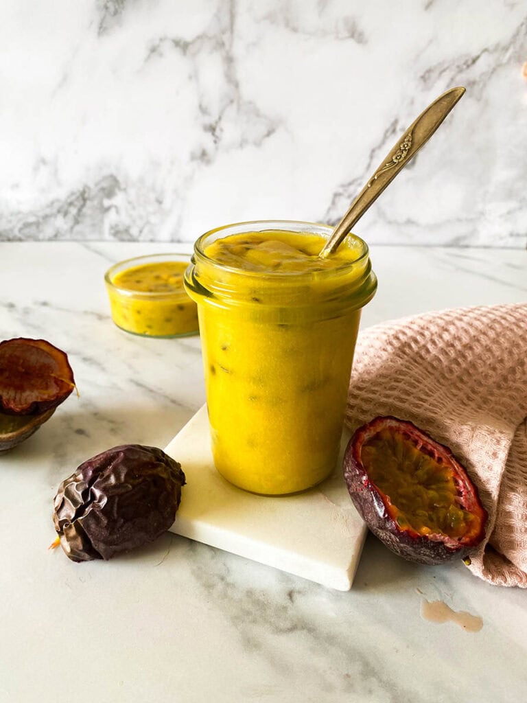 Passion Fruit Curd: Easy & Delicious 1 passion fruit curd jar on a marble background with a vintage spoon, with fresh passion fruits over a pink napkin