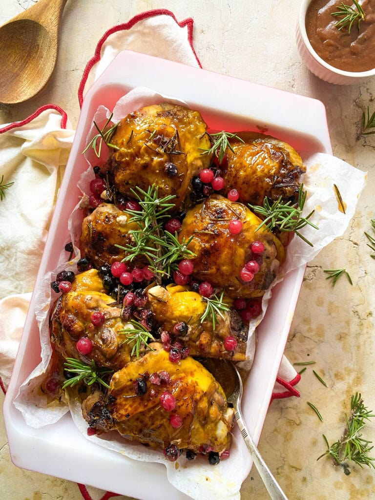 Cranberry Rosemary Chicken: The Delicious Christmas Feast 1 cranberry rosemary chicken in a baking tray over a red rimmed napkin, with a spoon and some fresh rosemary scattered around
