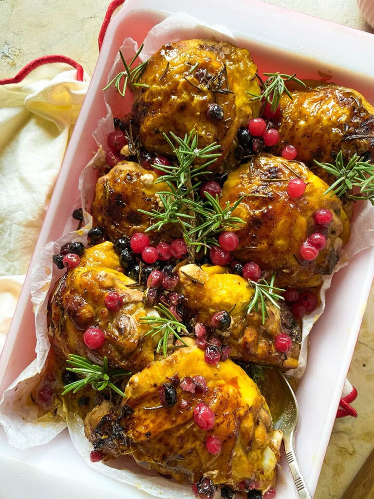Cranberry Rosemary Chicken: The Delicious Christmas Feast 2 cranberry rosemary chicken in a baking tray over a red rimmed napkin, with a spoon and some fresh rosemary scattered around