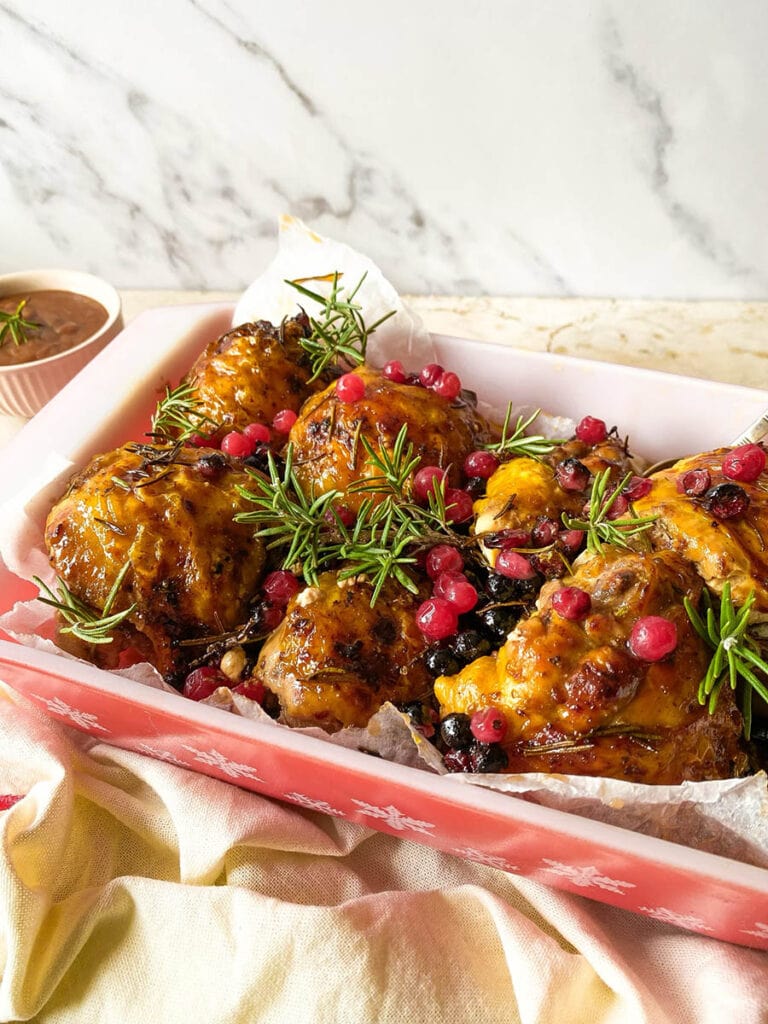 Cranberry Rosemary Chicken: The Delicious Christmas Feast 3 cranberry rosemary chicken in a baking tray over a red rimmed napkin, with a spoon and some fresh rosemary scattered around