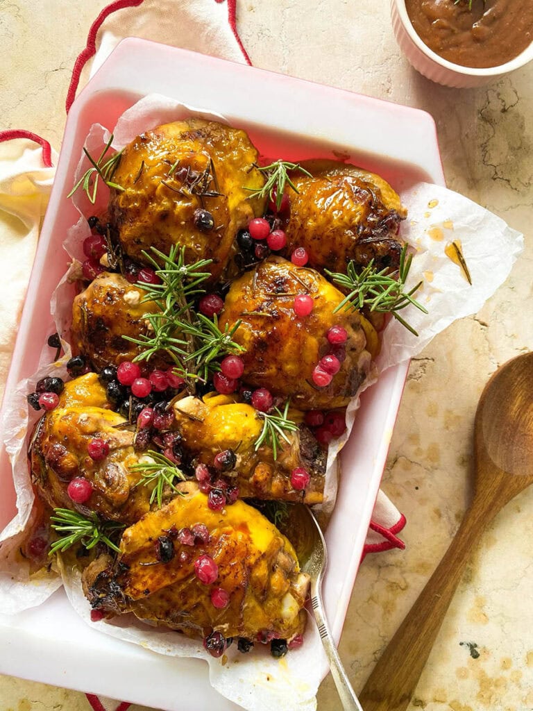 Cranberry Rosemary Chicken: The Delicious Christmas Feast 4 cranberry rosemary chicken in a baking tray over a red rimmed napkin, with a spoon and some fresh rosemary scattered around