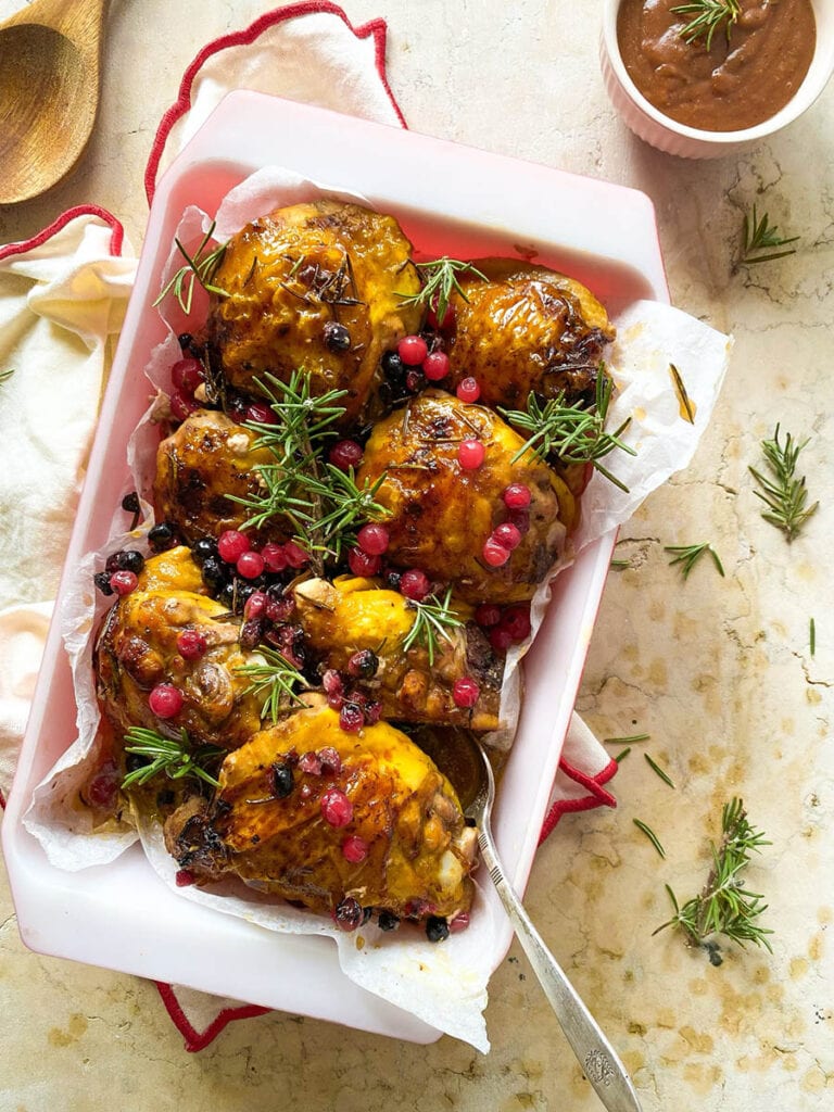 Cranberry Rosemary Chicken: The Delicious Christmas Feast 6 cranberry rosemary chicken in a baking tray over a red rimmed napkin, with a spoon and some fresh rosemary scattered around